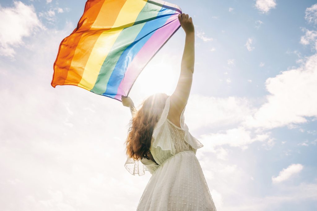 Frau in weißem Kleid streckt eine wehende Pride-Flagge Richtung Himmel