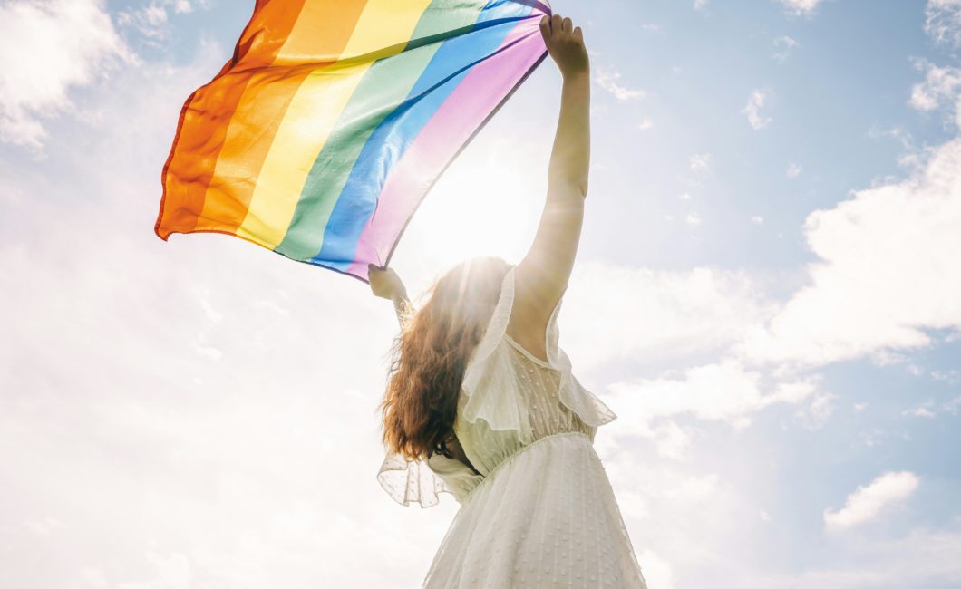 Frau in weißem Kleid streckt eine wehende Pride-Flagge Richtung Himmel