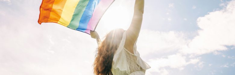 Frau in weißem Kleid streckt eine wehende Pride-Flagge Richtung Himmel